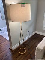 Full view of the gold standing floor lamp plugged in next to a wall and wooden floor with a large beige lampshade.