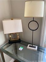 Photo showing two table lamps on a glass top side table: one with a rectangular gold-tone base and white shade, the other with a black stand featuring a circular ring and a white rectangular base with cream cylindrical shade.