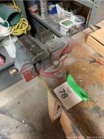 Full view of the mounted bench vice grip from the side showing the base attached to the wooden workbench with some tools and equipment in the background.