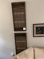 Photo showing two identical rectangular wood wall art panels mounted vertically on a wall above a couch. Both panels have a dark wood lattice grid design within a wooden frame.