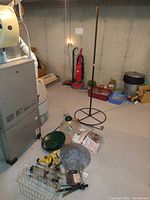 Photo shows basement storage area with various bird feeding equipment arranged on floor near furnace unit, including tall standing feeder pole, transparent dome squirrel baffler, metal basket, and multiple hanging feeders.