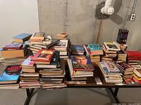 Wide shot showing multiple stacks of assorted books placed on a wooden table in a basement, featuring a variety of genres and formats.