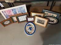 Multiple framed prints laid on floor and a ceramic decorative plaque in front showing a woman holding an umbrella, dressed in blue and white with cityscape background.