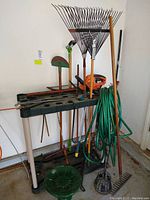 Garden tools and bird bath displayed on black plastic rack in garage corner. Items include rakes, hedge trimmer, hand saw, hose, and bird bath bowl.