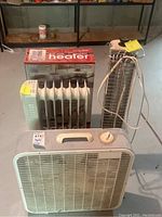 Front view of the three devices: Lasko electric fan, Comfort Zone boxed radiator heater, Boekamp vertical heater with power cord.