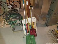 Hose organizer holding green garden hose beside three shovels, squeegee, and boxed heated birdbath in basement setting.