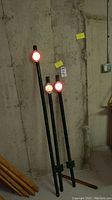 Three metal yard stakes with red round reflectors on top, standing against a concrete wall.