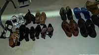 Photo showing two rows of mixed men's and women's shoes including sandals, casual leather shoes, rubber galoshes, and boots, arranged on a floor surface.