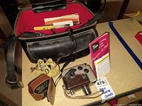 Vintage movie camera and accessories arranged near black leather camera case with red interior lining. Items include metal body Revere 8 B-61, Weston exposure meter, lenses in leather pouches, and instruction manuals.