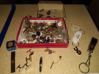 Wide view of assorted costume pins, cufflinks, tie clasps, and keychains on a flat surface with some items in a box and some outside.