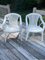 Two white plastic outdoor chairs positioned side by side on a wooden deck surface, showing their overall shape and condition.
