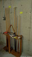 Wide view of six pool cues standing in wooden rack, pool accessories and instruction books on rack surface in basement setting.
