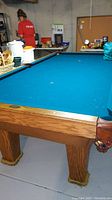 Angle view showing full length of blue felt playing surface, oak legs with felt pads, and side wooden rails with pockets.