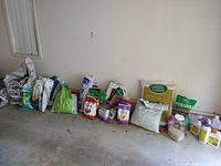 Photo showing a collection of bags of potting soil, peat moss, cedar mulch, pine spruce mulch, compost cow manure, and various gardening mixes lined up against a garage wall. Several bags are unopened while some are partially opened.