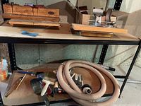 Photo showing electrical cords, multiple paint brushes in various sizes, and a coiled flexible grey hose with connectors, placed on a metal shelf.