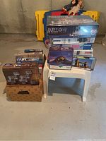Boxes of assorted jigsaw puzzles stacked on and around a white plastic table in basement setting.