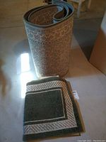 Rolled up "Ancient Garden" runner rug and two folded green and beige throw rugs on a basement floor surface, showing the pattern and condition of the rugs.