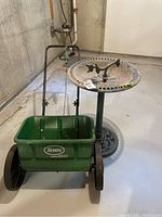 Photo showing green Scotts Accugreen 2000 seeder with black wheels and handle next to green plastic bird bath with pedestal base and decorative top with birds.