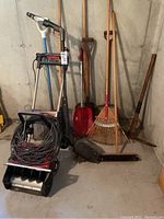 Photo of all the yard tools and power tools together in a basement corner including snow thrower, edger, rakes, shovels, broom, and pickaxe
