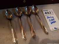 Four sterling silver spoons arranged in a row on a flat surface with a MaxSold auction tag nearby.