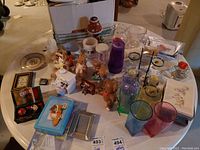 Assorted candle votives, figurines, mugs, plastic cups, greeting cards, and miscellaneous small items arranged on a round white table.