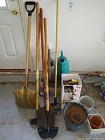 Photo showing garden tools including three shovels with wooden handles, plastic watering cans in teal and gray, various planter pots, a Chapin home and garden sprayer, and a caddy wagon stored in a garage environment.