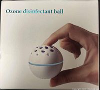 Front view of the white ozone disinfectant ball held between two fingers showing small ventilation holes on top and light blue ring around middle section.