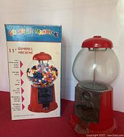Front view of the gumball machine next to its box showing colorful gumballs in globe, metal coin mechanism, and box features.