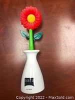 Front view of the white holder and flower-shaped sink brush with red bristles, green stem, and yellow center.