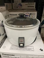 Panasonic white automatic rice cooker with stainless steel lid on original box, front view showing switch and indicator light.