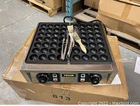 Front angle view of the takoyaki maker placed on a cardboard box with three grilling brushes and a whisk on top.