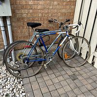 Two adult size bicycles, one Ascent silver frame and one Supercycle blue frame, leaning against a brick wall and fence on stone pavement.