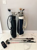 Two upright golf bags filled with golf clubs and additional clubs lying on floor with a club head cover.
