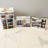 Photo of three puzzle boxes arranged side by side on the floor. The boxes are labeled 'Puzzlers 8 Collection', 'Puzzlers Collection 4', and 'Thomas Kinkade 10'.