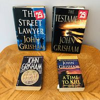 Four John Grisham novels grouped on wooden table: two hardcover (The Street Lawyer, The Testament) with discount stickers, two paperback (The Client, A Time to Kill) showing wear and creases.