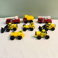 Photo showing all 8 Tonka toy trucks arranged in two rows on a white surface with a plain background. Includes various types: fire trucks, dump truck, and construction vehicles.