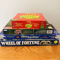 Pile of four board games visible from top and side; Apples to Apples Party Box at top, WhoVille-opoly and Wheel of Fortune below.