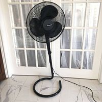 Front view of black floor fan with metal grille and three blades, standing on circular base indoors against white doors.