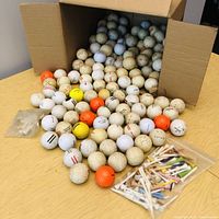 Photo of a large open box with over 160 used golf balls spilling out, alongside a small bag of assorted wooden golf tees.
