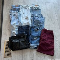 Grouped view of six pairs of jeans, black faux leather shorts, and a burgundy skirt laid on a wood floor