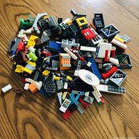 Pile of mixed LEGO pieces showing various bricks, plates, window frames, wheels, and specialty parts spread on a wooden floor.
