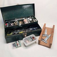 Green metal toolbox open showing hand tools and hardware inside, wooden mitre box and plastic container with hooks in front