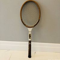 Front view of vintage wood Dunlop tennis racket showing the head, strings, and handle wrapped in brown grip tape against a white tiled floor and grey wall.