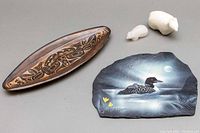 Photo showing wooden elongated dish with Haida style carvings, and a painted clay plate featuring a loon on water under moonlight, signed Haida Indigenous art style.