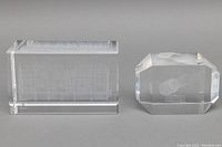 Two crystal paperweights side by side on gray background. Left is rectangular with 3D satellite inside; right is smaller multifaceted with 3D shuttle.