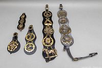 Five vintage black leather straps arranged horizontally, each with multiple polished brass horse bridle medallions of varying sizes, showing an assortment of horse-related designs including crowns, horseshoes, and horses.