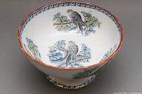 Oval white porcelain footed bowl with hand painted colorful birds and tree branches visible inside and out, highlighted with an orange-red rim and blue floral border on foot.