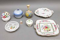 Full view of all 8 porcelain items laid out on gray background including cake plates, bowls, mini bell, floral arrangements, and pitcher
