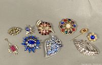 Eight assorted vintage costume jewelry pieces including brooches, pins, and a pendant arranged on a flat surface for overview.