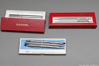 Three boxed sets of pens and mechanical pencils on a gray background. The Sheaffer sets are in red boxes and the Cross set is in a white and blue box.